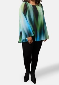 Top lungo fluido e trasparente in gradiente blu e verde con scollatura rotonda, abbinato a pantaloni slim-fit neri e décolleté nere a punta.