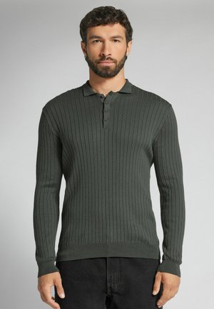 Mann mit dunklem lockigem Haar und Bart, der ein dunkelgrünes, geripptes Langarm-Poloshirt und schwarze Hose trägt, steht vor einfachem Hintergrund.