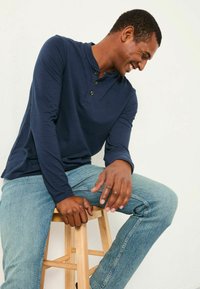 Marineblaue Langarm-Henley-Bluse mit drei Knöpfen, getragen mit hellblauen Jeans. Das Shirt hat ein dezentes Logo auf der Brust.