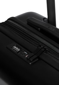 Valigia rigida nera con finitura opaca, chiusura con zip e lucchetto a combinazione. Presenta un manico retrattile e dettagli in zip testurizzati.