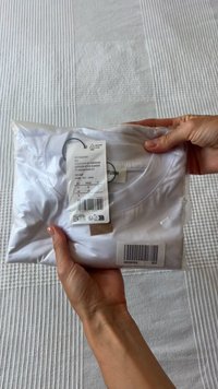 Camiseta de algodón blanca, cuidadosamente empaquetada en una funda de plástico transparente, con una etiqueta que incluye detalles de tamaño, SKU y código de barras.