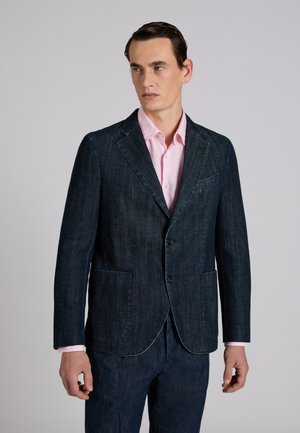 Mężczyzna noszący ciemny denimowy blezer i pasujące spodnie, na jasnoróżowej koszuli na guziki, stojący na jednolitym tle.