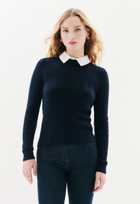 Marineblaue Strickjacke mit einem weißen Hemd darunter. Sie hat lange Ärmel, einen gerippten Saum und einen strukturierten Stoff.