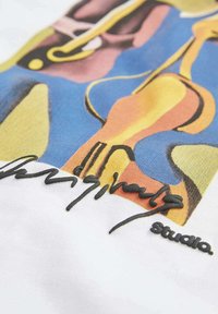 Camiseta de algodón blanca con un estampado abstracto colorido en tonos amarillos, azules y rosas. Incluye una firma bordada en negro y etiqueta "Studio".