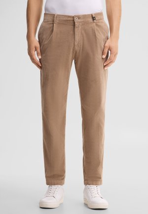 Mann trägt beige Cordhosen und weiße Sneakers, steht vor einem einfarbigen hellen Hintergrund.
