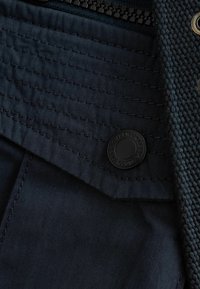 Marineblaue Stoff mit strukturiertem Gewebe, vertikalen Nähten und einem schwarzen runden Logo-Knopf als Akzent. Reißverschlussteil am oberen Ende sichtbar.