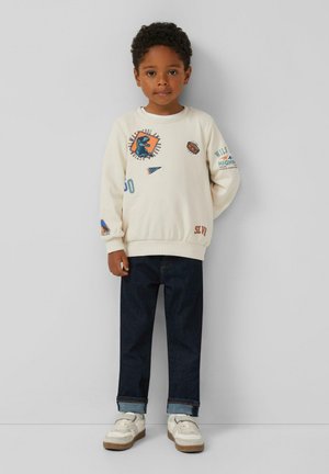 Cremfarbener Sweatshirt mit Grafikdruck, Rundhalsausschnitt und langen Ärmeln, kombiniert mit dunkelblauen Jeans und weißen Sneakers.