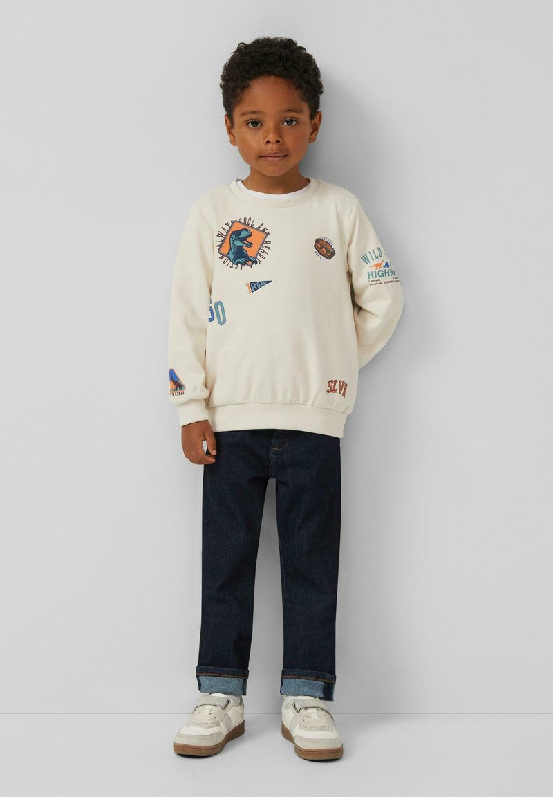Cremfarbener Sweatshirt mit Grafikdruck, Rundhalsausschnitt und langen Ärmeln, kombiniert mit dunkelblauen Jeans und weißen Sneakers.