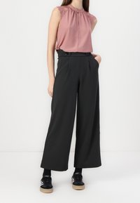 Femme portant un chemisier sans manches rose poudré avec des épaules en dentelle, un pantalon noir taille haute à jambes larges, des chaussettes noires et des baskets noires à rayures blanches.
