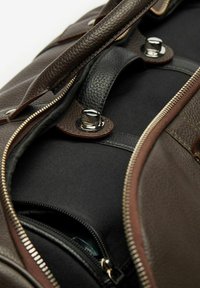 Ruskea nahkainen duffel-laukku mustalla kankaalla, jossa on metalliset osat, kaksoist vetoketjut ja kontrastivärinen ommel reunoilla.