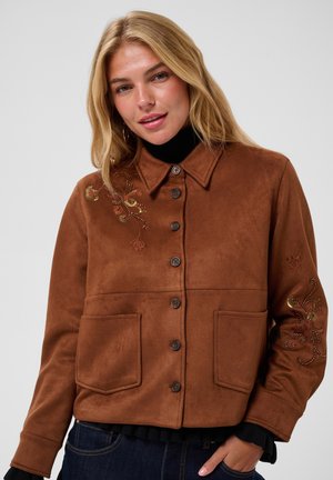 Blonde vrouw met een bruin jasje met knopen en bloemrijke borduursels op de schouder en mouw, staand met één hand in de zak.