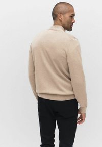 Beżowy sweter dzianinowy z okrągłym dekoltem i ściągaczami, noszony z czarnymi spodniami, ukazujący prostą, gładką fakturę i czysty design.