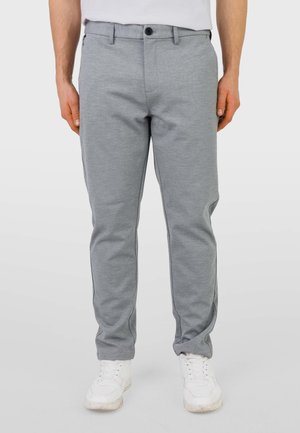 Uomo con pantaloni grigio chiaro aderenti e scarpe da ginnastica bianche, in piedi con le mani lungo i fianchi davanti a uno sfondo semplice.