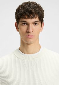 Maglione bianco a coste, con collo rotondo, caratterizzato da dettagli testurizzati e una vestibilità aderente, indossato sopra una t-shirt bianca con collo a giro.