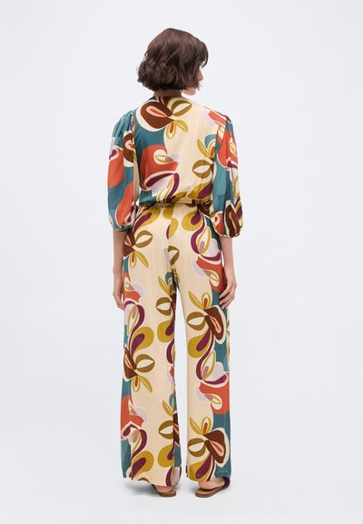 Γυναίκα με κοντά καστανά μαλλιά, που φοράει πολύχρωμο πουκάμισο με σχέδια και παντελόνι με φαρδιά μπατζάκια, στέκεται γυρισμένη προς τα πίσω σε απλό φόντο.