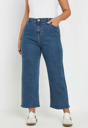 High-waisted, wijde blauwe denim jeans met een gladde textuur, gouden knoopjes, en voorzakken, gecombineerd met witte sneakers.