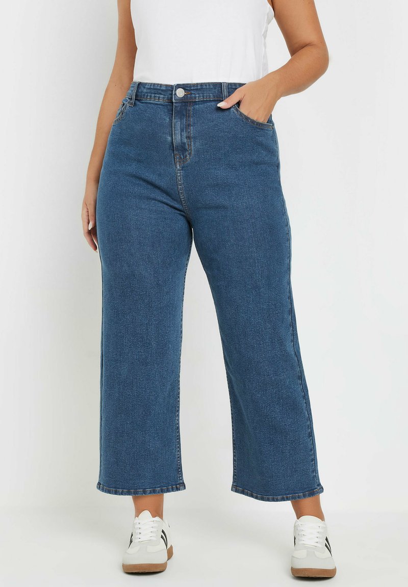 High-waisted, wijde blauwe denim jeans met een gladde textuur, gouden knoopjes, en voorzakken, gecombineerd met witte sneakers.