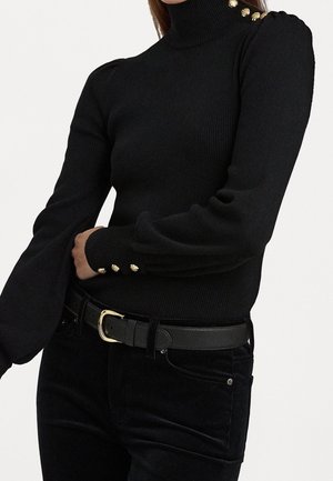 Femme portant un pull à col roulé côtelé noir avec des boutons dorés sur l'épaule et les poignets, associé à un pantalon noir et une ceinture noire avec une boucle dorée.