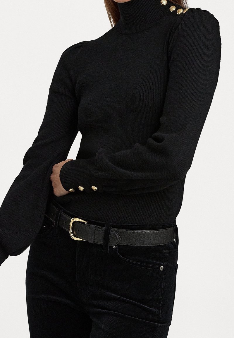 Femme portant un pull à col roulé côtelé noir avec des boutons dorés sur l'épaule et les poignets, associé à un pantalon noir et une ceinture noire avec une boucle dorée.
