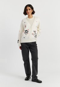 Elfenbeinfarbiger Cardigan mit schwarzen und roten Illustrationen, ausgestattet mit einem V-Ausschnitt, Frontknöpfen und gerippten Bündchen. Kombiniert mit grauen Jeans und schwarzen Schuhen.