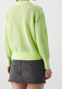 Maglione verde lavorato a maglia con un motivo a trama larga, polsini e orlo a coste. Indossato sopra una mini gonna in denim scuro con orli sfrangiati.