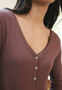 Maglione pullover a V in colore malva con texture a coste, decorato con piccoli bottoni lungo la parte frontale e un bordo ondulato sul colletto.