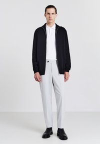 Schwarze Zip-Jacke über einem weißen Poloshirt, kombiniert mit hellgrauen, maßgeschneiderten Hosen. Das Model trägt schwarze Stiefeletten. Glatter Stoff und taillierter Schnitt.