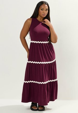 Kvinde med langt mørkt hår iført en ærmeløs lilla maxi-kjole med hvide bølgede bånd, stående med den ene hånd nær ansigtet.