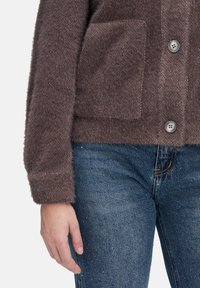 Brun tekstureret fleecejakke med en frontlomme og knaplukning, parret med blå denimjeans med en lys vask og subtile detaljer.