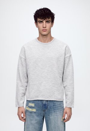 Ung mand med krøllet mørkt hår iført en lysegrå langærmet trøje og slidte blå jeans mod en ensfarvet baggrund.