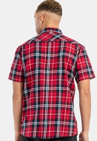 Muž s krátkými kudrnatými vlasy, oblečený v červené, černé a bílé kostkované košili s krátkým rukávem, zobrazený zezadu na jednobarevném pozadí.