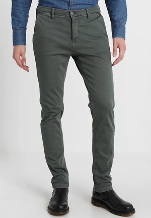 Homme portant un pantalon slim vert olive, des bottes en cuir noires et une chemise en denim bleu, debout devant un fond uni.