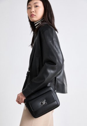 Black leather crossbody bag with silver hardware, rectangular shape, front flap, and embossed logo. Textured finish against a model in a black jacket.