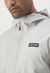 Giacca beige chiaro con cappuccio, dotata di un logo Calvin Klein nero sul lato sinistro del petto, texture liscia e dettagli con chiusura a zip.