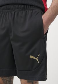 Svarta sportshorts i lättviktsmaterial, med elastisk midja, sidofickor och en guld Puma-logotyp på vänster ben.