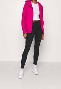 Ljusrosa zip-up hoodie med huva, kombinerad med svarta högmidjade leggings och vita träningsskor, mot en slät bakgrund.