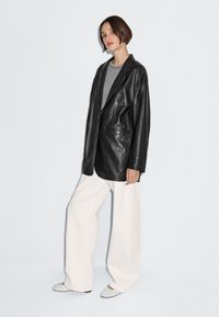 Černý kožený blejzr s nadměrným střihem, zapínáním na jeden knoflík a dvěma předními kapsami, nošený přes šedý top a doplněný bílými širokými kalhotami.