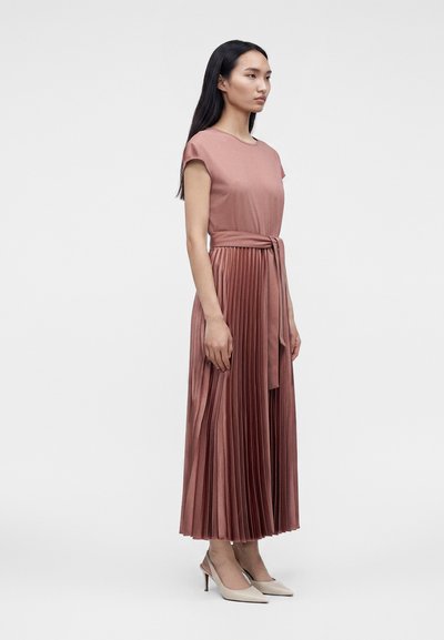 Femme aux longs cheveux foncés, debout de profil, portant une robe plissée rose poudré avec une ceinture à la taille et des talons pointus beiges.