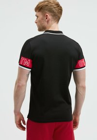 Polo nero con accenti rossi e bianchi sulle maniche, caratterizzato dalle lettere "TM" e "01". Tessuto texture, vestibilità standard, colletto semplice.