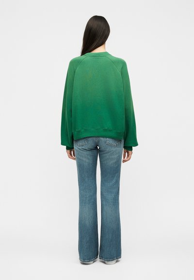 Γυναίκα με μακριά σκούρα μαλλιά που φοράει πράσινο φούτερ και μπλε καμπάνα τζιν, στέκεται με την πλάτη γυρισμένη σε ένα απλό λευκό φόντο.