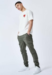 T-shirt bianco a maniche corte con una grafica di un cuore rosso, abbinato a pantaloni cargo oliva con più tasche e sneaker bianche.