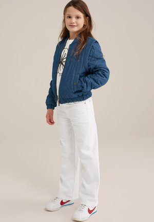 Veste bomber matelassée bleue avec des poignets côtelés, portée sur un t-shirt graphique blanc, associée à un jean blanc et des baskets à accents rouges.