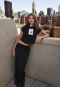 Zwarte cropped t-shirt met grafische print, gecombineerd met een lange zwarte rok. Achtergrond van een stedelijk dak met de skyline van de stad.