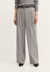 Hellegraue, weite Hose aus strukturiertem Stoff, mit vorderen Falten, einem schwarzen Gürtel mit einer Schnalle aus goldfarbenem Metall und spitzen schwarzen High Heels.