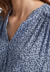 Väljande blå blus med v-ringning, svart blommönster och texturerad tyg. Rynkade axlar ger en avslappnad passform.