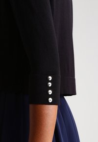 Svart stickad topp med trekvartsärmar och silverknappar på manschetten, matchad med en mörkblå kjol.