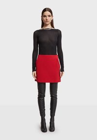 Rode A-lijn rok gecombineerd met een zwarte transparante top met lange mouwen. Model draagt zwarte laarzen tot boven de knie, met een zilveren ketting zichtbaar.