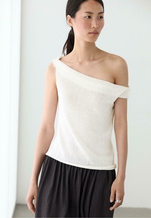 Kvinde iført en hvid strikbluse med bar skuldre og løse sorte bukser, der står mod en ensfarvet hvid baggrund med afslappede hænder.