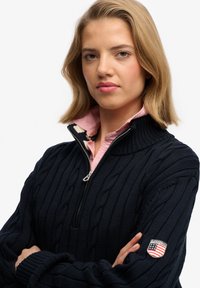 Maglione blu navy a maglia intrecciata con colletto alto e chiusura con zip. Presenta una camicia rosa con colletto sotto e una toppa sulla manica sinistra.