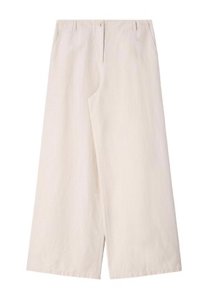 Persoon draagt crèmekleurige broek met wijde pijpen, bijpassend gilet, gouden ringen en met studs versierde bruine instapschoenen, staand voor een beige achtergrond.